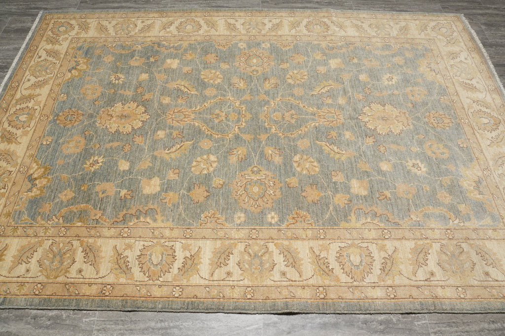 A rectangular area rug with a traditional floral pattern, featuring a muted blue-gray center field and warm beige borders, laid out on a gray floor.