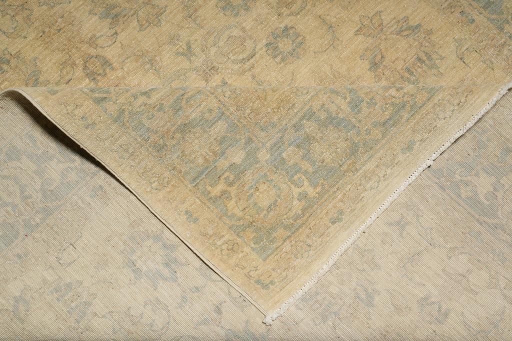 A close-up of an area rug with a faded traditional floral pattern in beige and muted blue-gray tones, showing the corner and fringed edge of the textile.