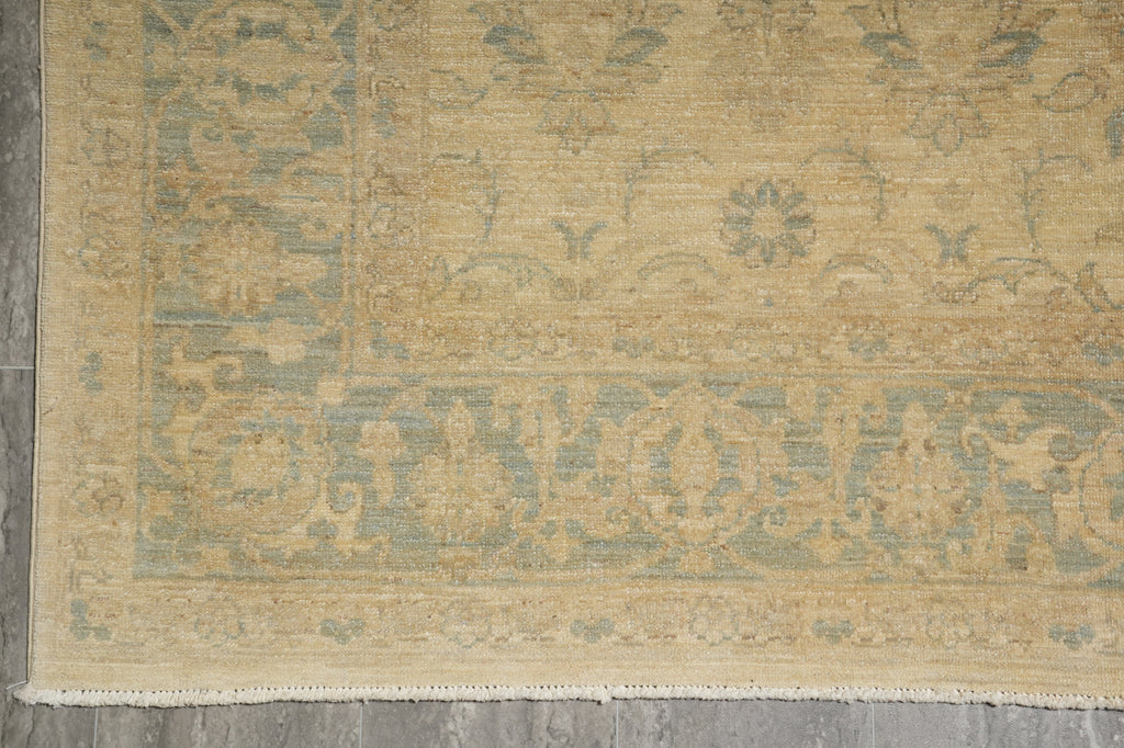 A close-up of a traditional area rug with a faded beige and muted blue-green floral pattern, laid out on a gray tiled floor.