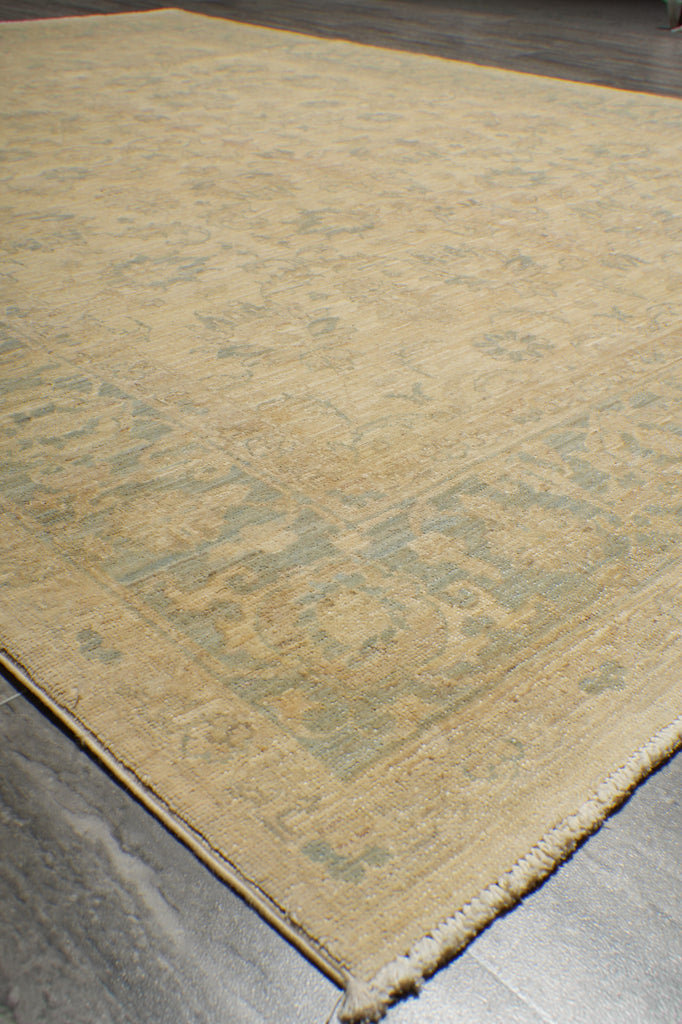 A close-up view of a beige area rug with a faded traditional floral pattern in muted blue-gray tones, shown on a dark gray floor.