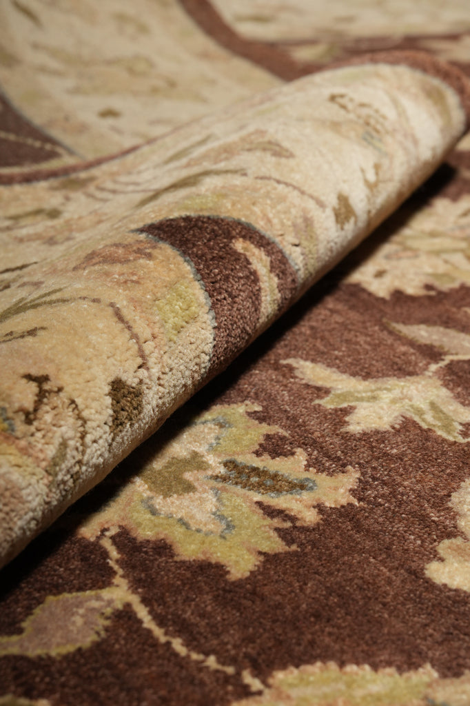 A close-up of a patterned rug or textile with a rolled section showing its texture and design. The color palette features warm brown, beige, cream, and muted olive-green tones in an ornate floral or damask-style motif.