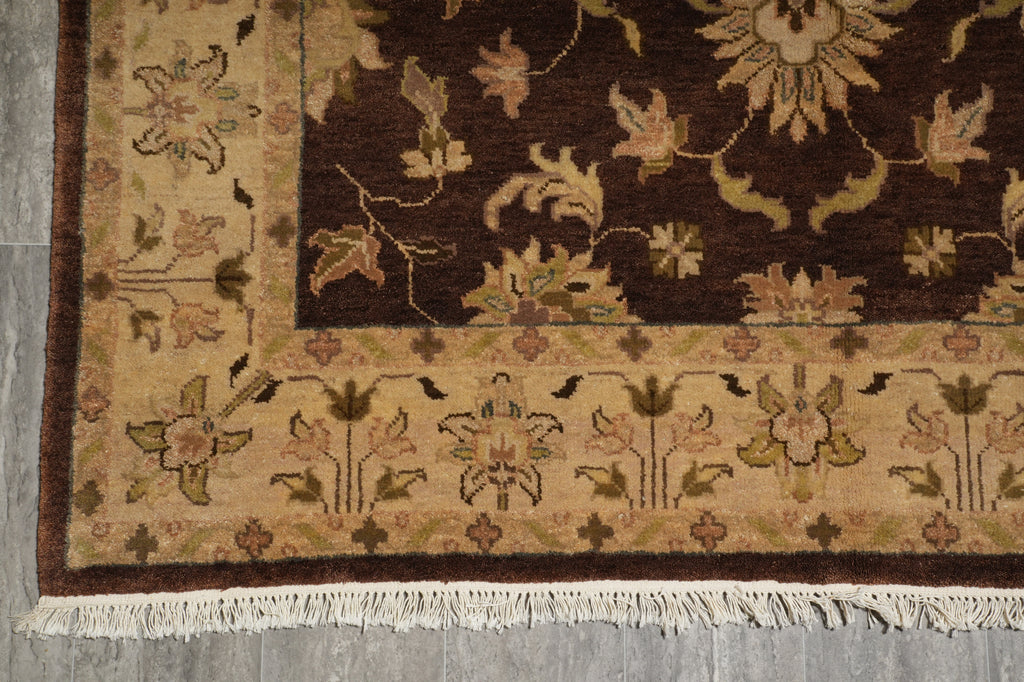 A close-up of a traditional patterned rug corner showing a dark brown field, beige border, floral motifs, and white fringe on a gray floor.
