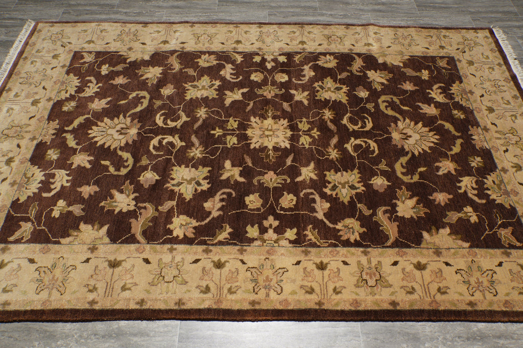 A rectangular area rug with an ornate floral and vine pattern, featuring a dark brown center field and beige borders, laid out on a gray tiled floor.