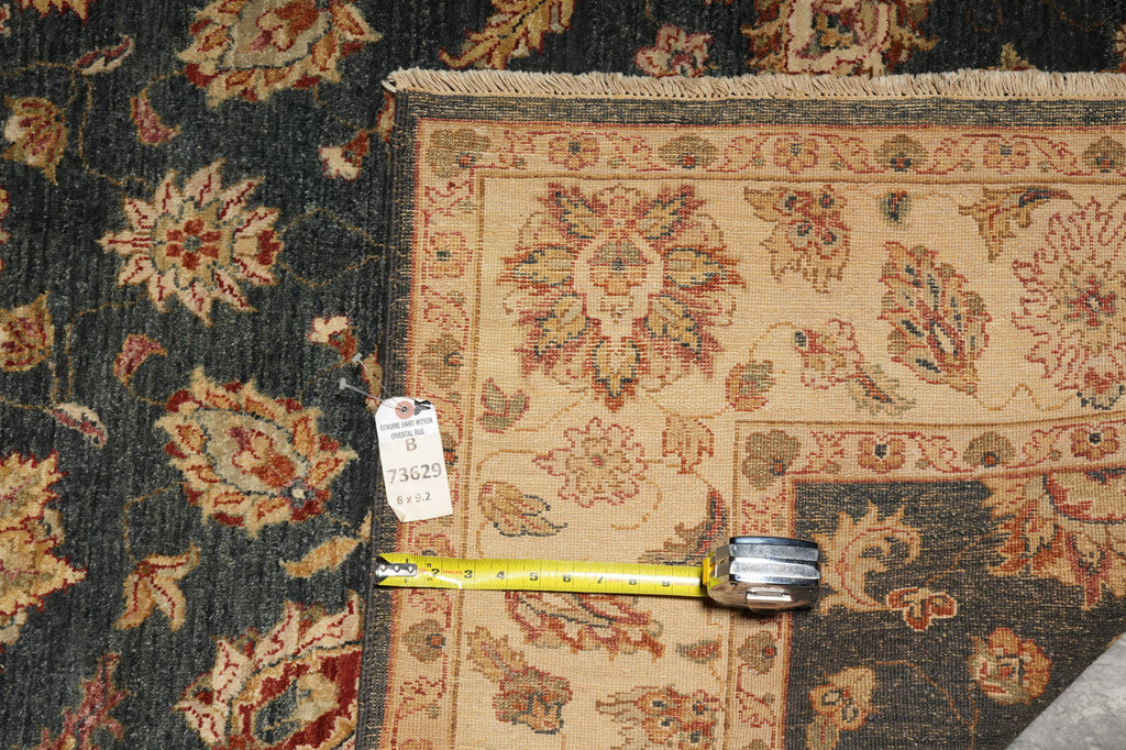 A close-up of a patterned area rug corner with a tape measure placed along the edge to show dimensions. The rug has a beige central field with floral motifs and a dark navy outer section with red and beige detailing.