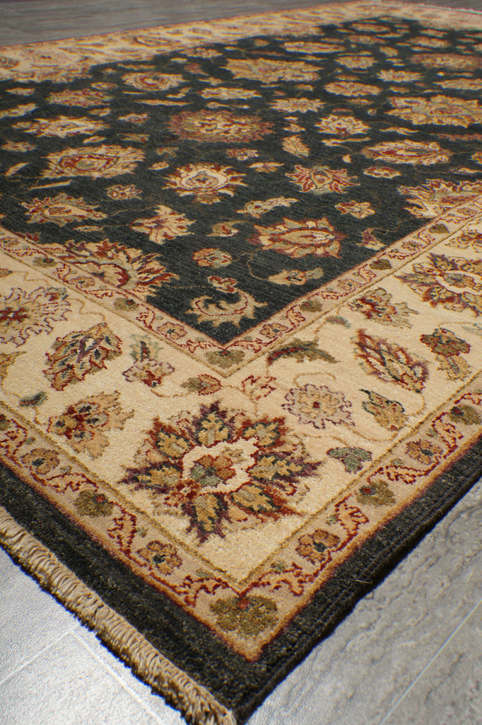A close-up of a traditional patterned area rug shown at an angle, featuring a dark navy blue field with floral motifs, a beige inner section, and a dark border.