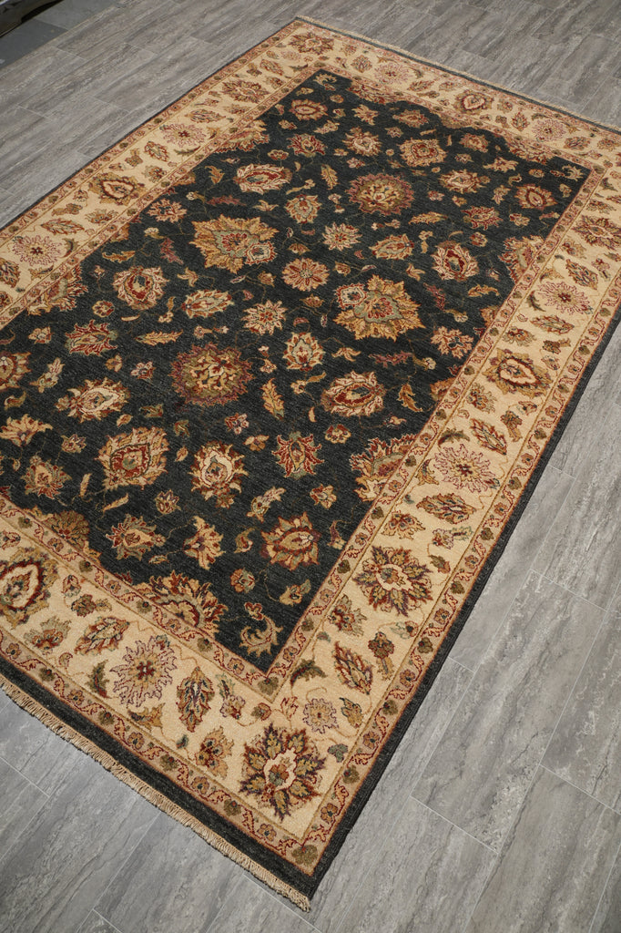 A rectangular area rug with an ornate floral pattern, featuring a dark charcoal-black center field, beige borders, and rust-orange accents, shown laid out on a gray wood-look floor.