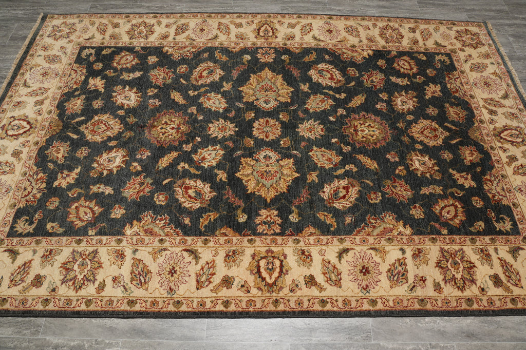 A rectangular area rug with an ornate traditional floral pattern, featuring a dark charcoal-black center field surrounded by beige borders on a gray wood floor.