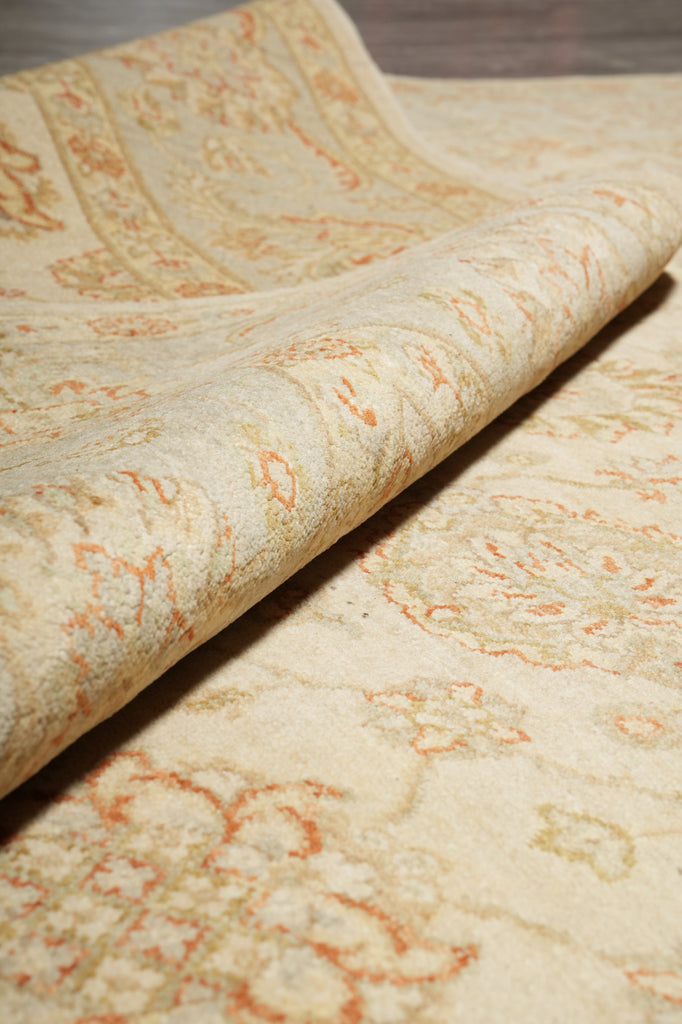 A close-up of a patterned rug with a rolled corner showing its texture and design. The rug has a warm cream base with intricate floral and scroll motifs in muted orange and light beige.