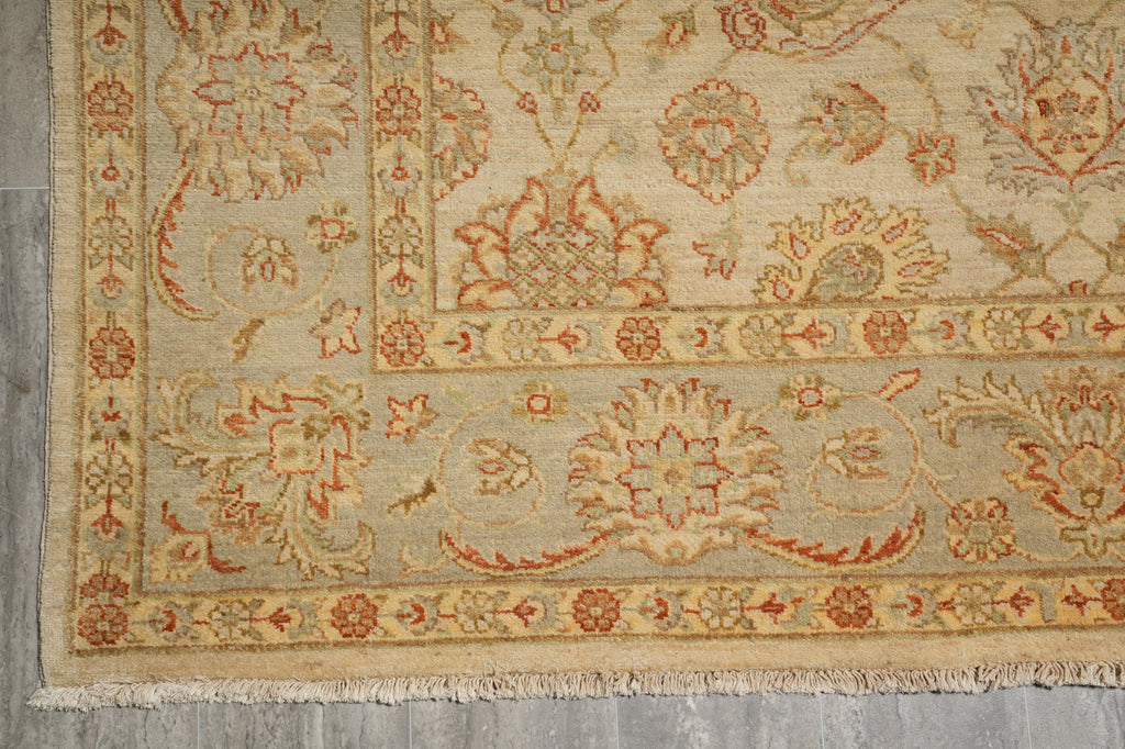 A close-up of a traditional patterned rug with a beige base and red, orange, and muted green floral motifs, showing the woven texture and fringe on a gray floor.