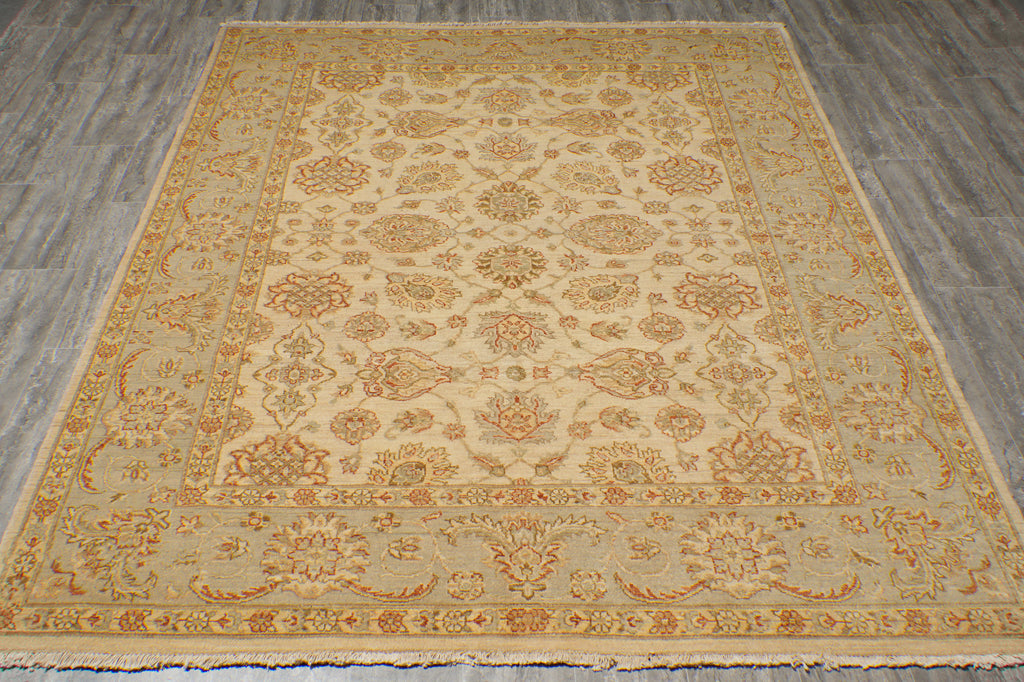 A rectangular area rug with an ornate traditional floral pattern, laid out on a gray wood-look floor. The rug features a warm beige center with a border of rust orange, tan, and muted green details.