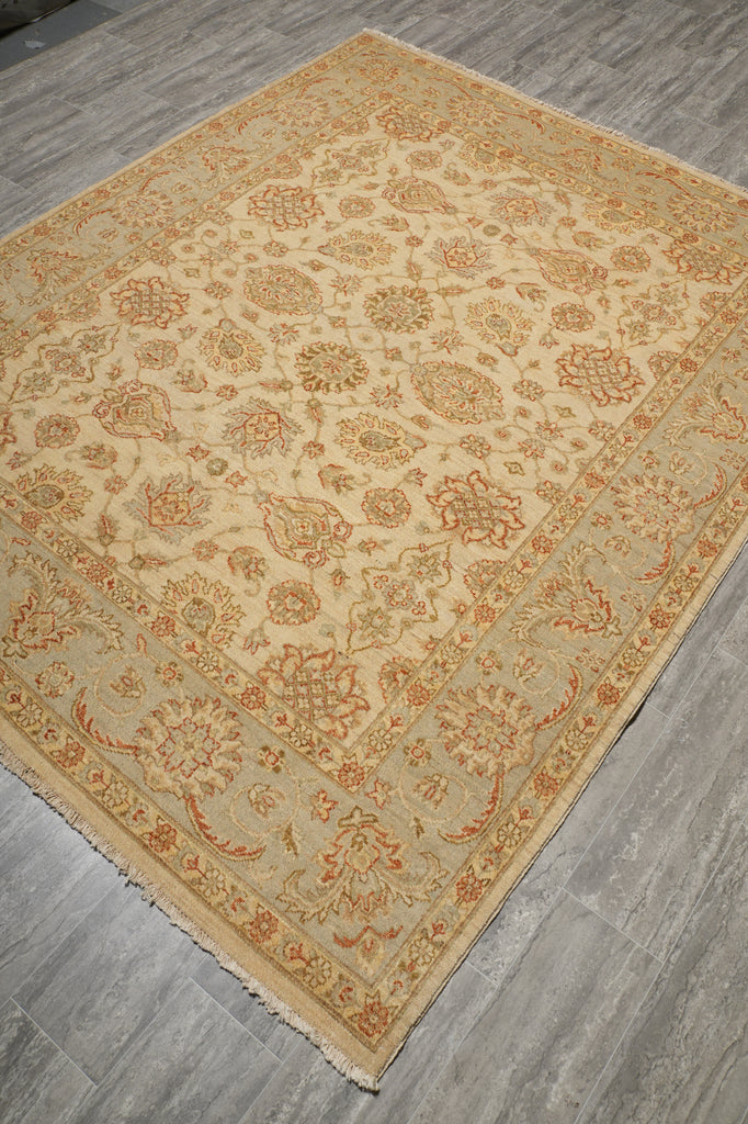 A rectangular area rug with an intricate floral and botanical pattern in beige, tan, and muted orange, laid out on a gray wood-look floor.