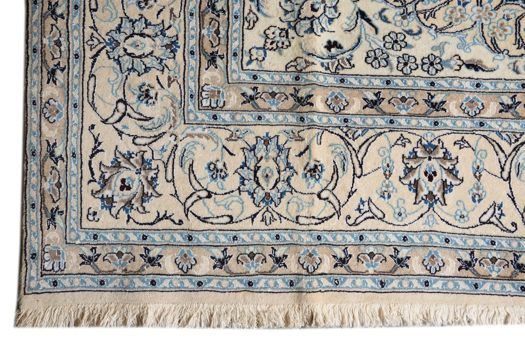 A close-up of an ornate area rug corner showing a cream base with intricate blue and dark brown floral motifs and fringed tassels.