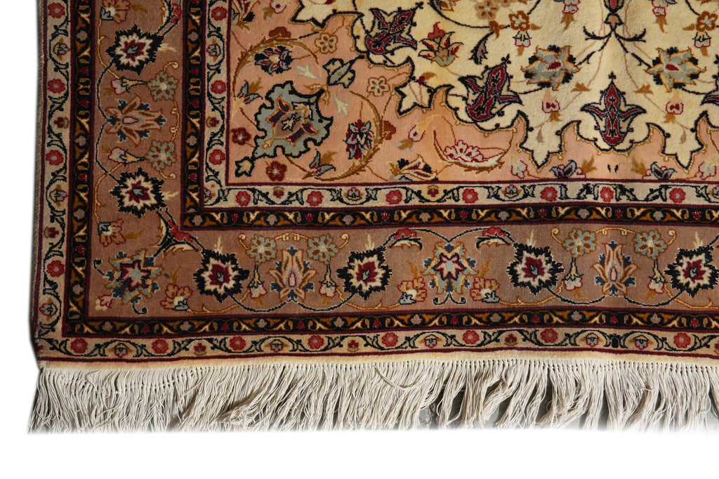A close-up of an ornate Persian-style area rug corner, showing a cream and tan field with a detailed floral pattern, a brown and red border, and a white fringe at the bottom.