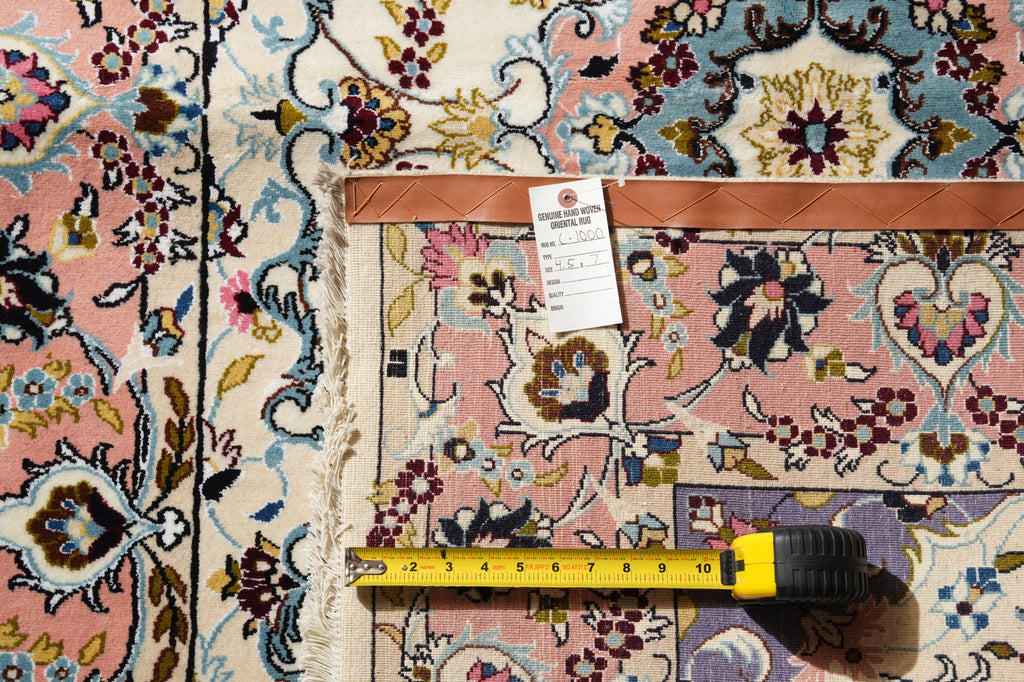 A close-up of a patterned area rug with a pink field, blue and beige motifs, and a brown leather border. A measuring tape and a product tag are placed on top of the rug to show scale and details.