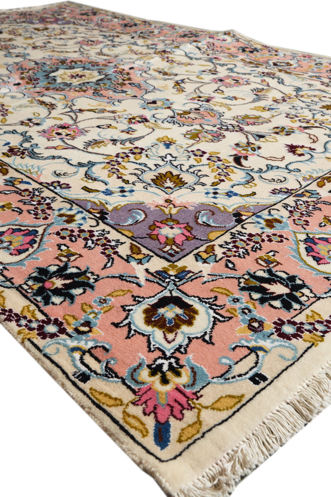 A close-up view of a traditional Persian-style area rug with a cream background, pink and blue floral motifs, and a purple central medallion, showing the detailed pile and fringe on one end.