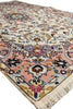 Load image into Gallery viewer, A close-up view of a traditional Persian-style area rug with a cream background, pink and blue floral motifs, and a purple central medallion, showing the detailed pile and fringe on one end.