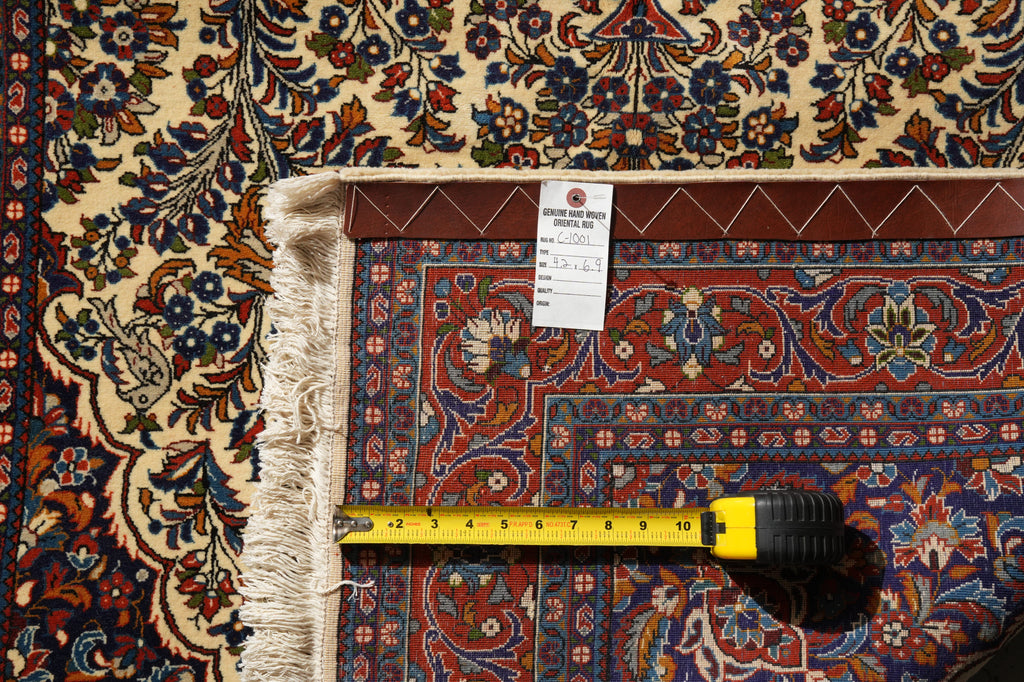 A close-up of an ornate patterned area rug with a cream fringe, a white size tag, and a yellow tape measure laid across it to show dimensions.