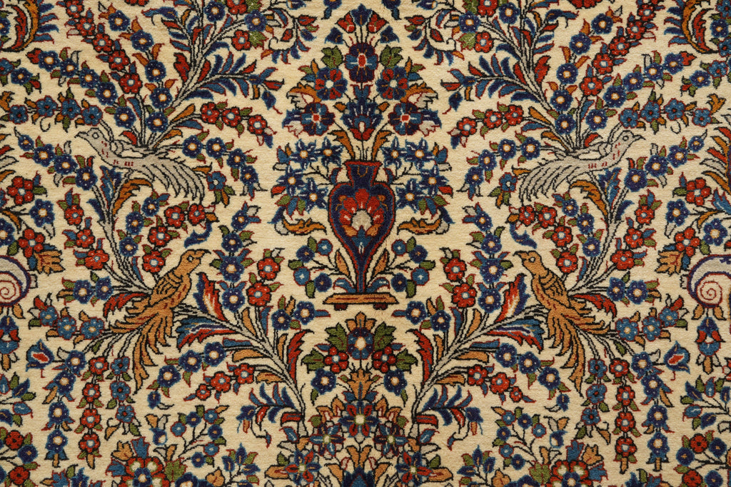 A close-up of an ornate patterned textile or rug featuring a cream background with dense floral motifs in blue, red, green, and gold, plus stylized birds and a central vase design.