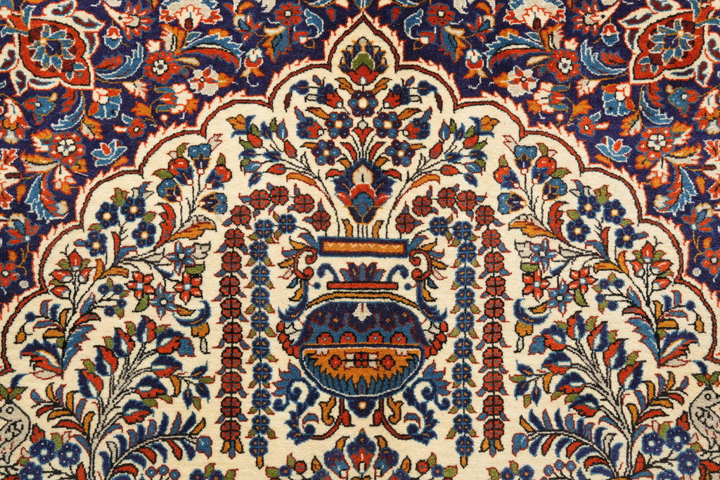 A close-up of an ornate traditional patterned rug featuring a central floral medallion and symmetrical scrolling motifs in cream, navy blue, rust red, and orange.