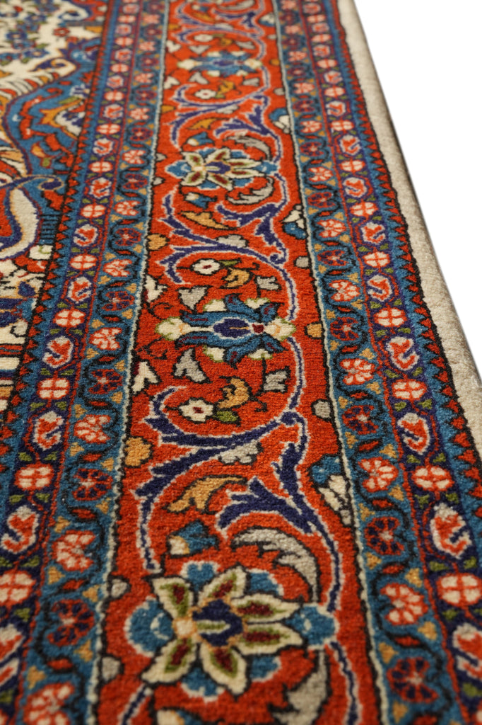 A close-up view of an ornate patterned area rug with a vibrant red center, detailed blue and beige floral motifs, and a cream-colored border.