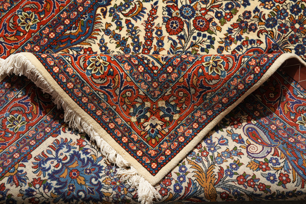 A close-up of a traditional Persian-style area rug with an intricate floral and paisley pattern in red, blue, and cream, showing the detailed weave and cream-colored fringe.