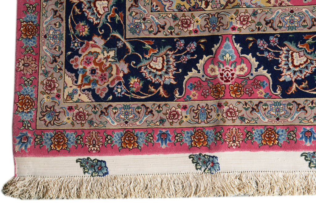 A close-up of a traditional oriental rug corner showing a dark navy blue field with ornate floral motifs, a pink border, beige inner bands, and cream fringe along the bottom edge.