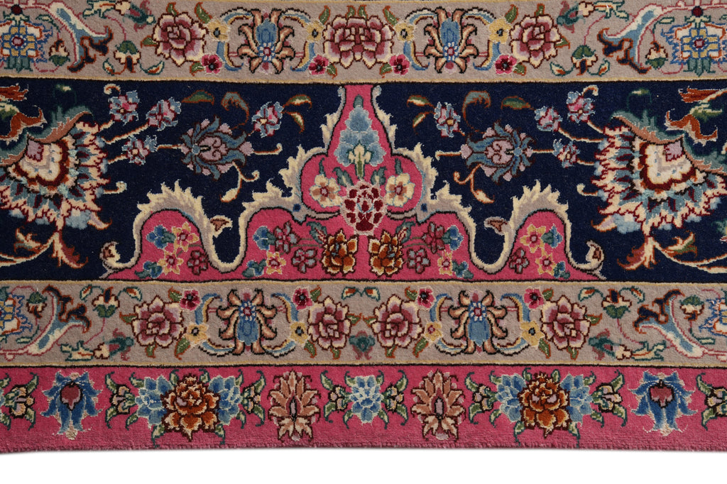A close-up of a traditional patterned rug featuring a navy blue central field, pink and beige borders, and floral motifs in blue, gold, and rust.