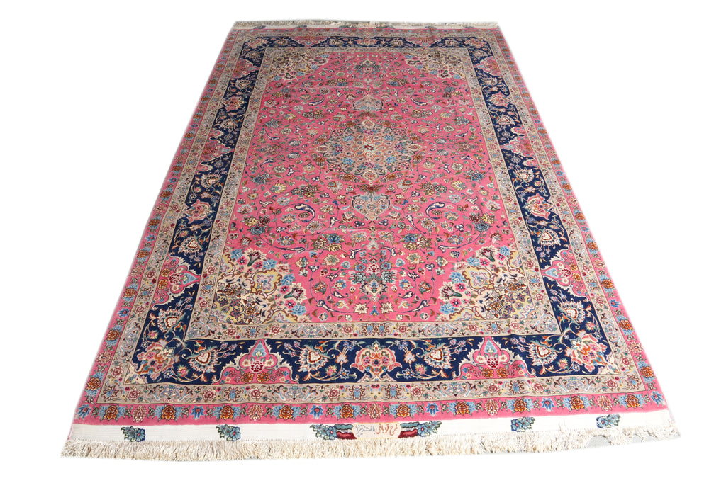 A traditional oriental area rug with a pink field, navy blue borders, and intricate floral motifs in beige, green, and blue, finished with cream fringe on the ends.