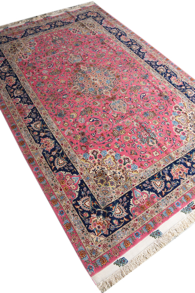 A traditional Persian-style area rug with a vibrant pink field, dark navy blue borders, and intricate floral motifs in beige, blue, and green, finished with cream fringe.