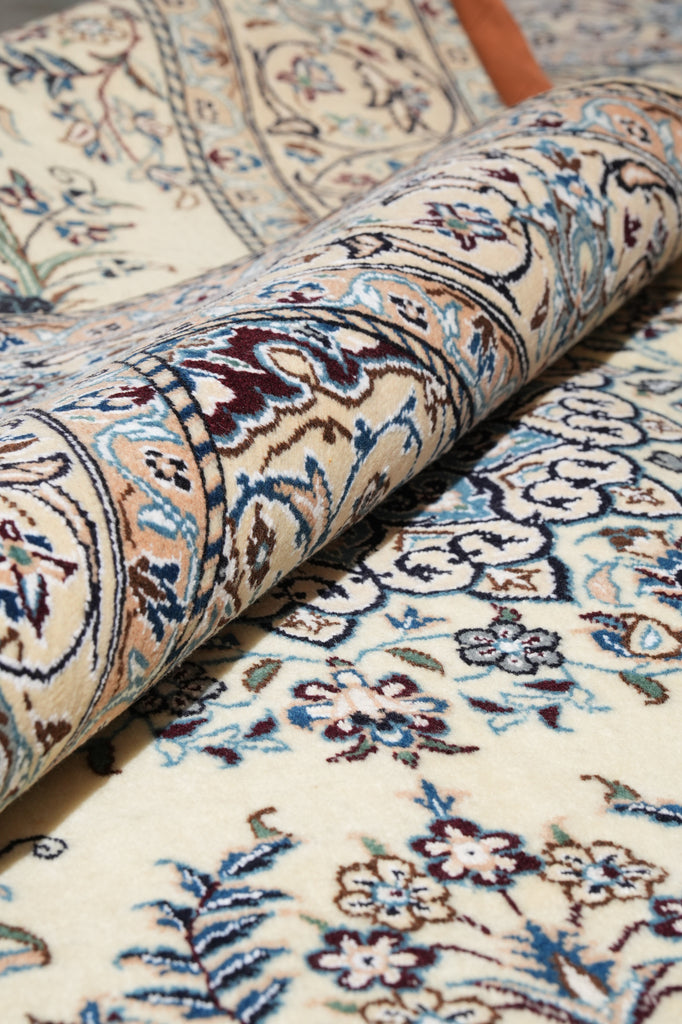 A close-up of a rolled section of an ornate patterned rug showing cream, blue, burgundy, and brown floral and paisley motifs.