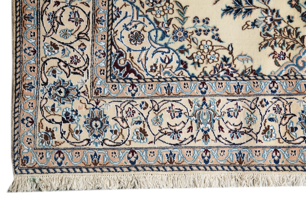 Close-up of an ornate area rug corner showing a cream base with intricate blue, brown, and beige floral and vine patterns and fringe along one edge.