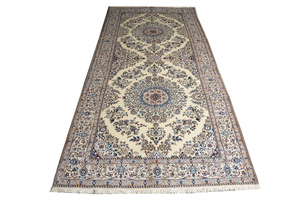 A long rectangular area rug with an intricate traditional Persian-style floral pattern, featuring cream, beige, blue, and brown tones, shown flat against a white background.