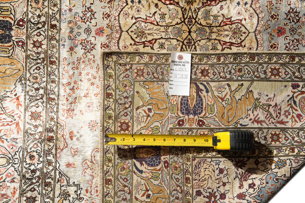 An ornate traditional patterned rug shown in a close-up flat lay, with a yellow tape measure laid across the center and a white product tag attached near the top. The rug has a cream base with red, blue, and brown floral and geometric motifs.