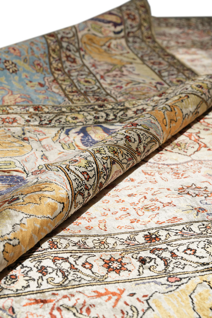 A close-up of a folded traditional patterned rug showing detailed floral and ornamental motifs in beige, cream, gold, blue, and rust tones.