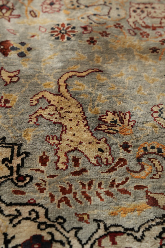 Close-up of an ornate rug or textile with a beige animal motif on a muted gray-beige background, surrounded by red, brown, and black floral and scroll patterns.