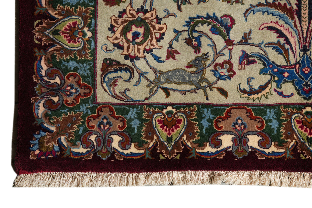 Close-up of a patterned area rug corner showing a cream field with a blue and green deer motif, ornate floral borders in burgundy, teal, and gold, and cream fringe along the bottom edge.