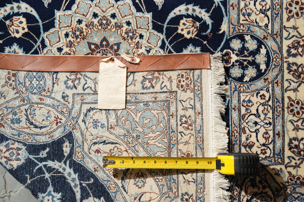 A close-up of an ornate patterned rug with cream, blue, and brown tones, showing a leather trim band, a blank tag, and a yellow tape measure laid across the fringe to indicate size.