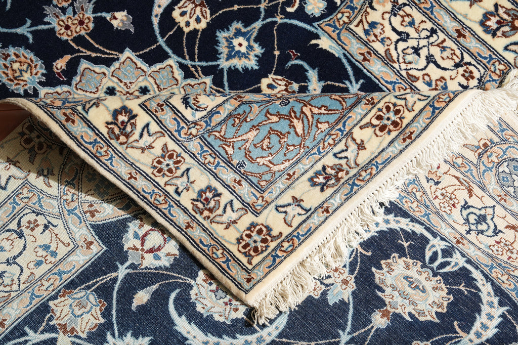 A close-up of an ornate Persian-style area rug with a navy blue field, cream borders, and light blue floral motifs, shown folded to reveal its cream fringed edge.