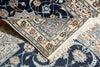 Load image into Gallery viewer, A close-up of an ornate Persian-style area rug with a navy blue field, cream borders, and light blue floral motifs, shown folded to reveal its cream fringed edge.
