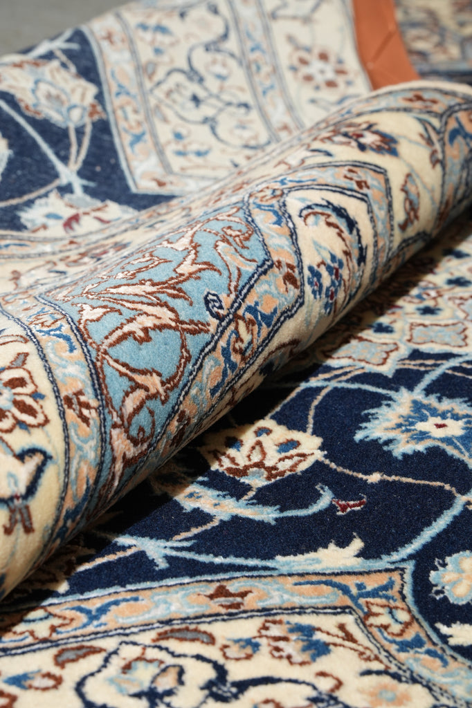 A close-up of a rolled area rug showing an intricate traditional pattern with navy blue, cream, light blue, and brown tones.