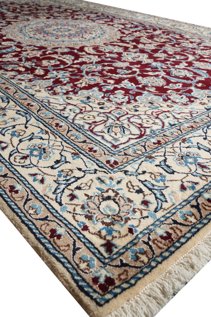 A close-up view of a traditional Persian-style area rug with a cream base, deep red central field, and blue and beige floral motifs, featuring a fringed edge.