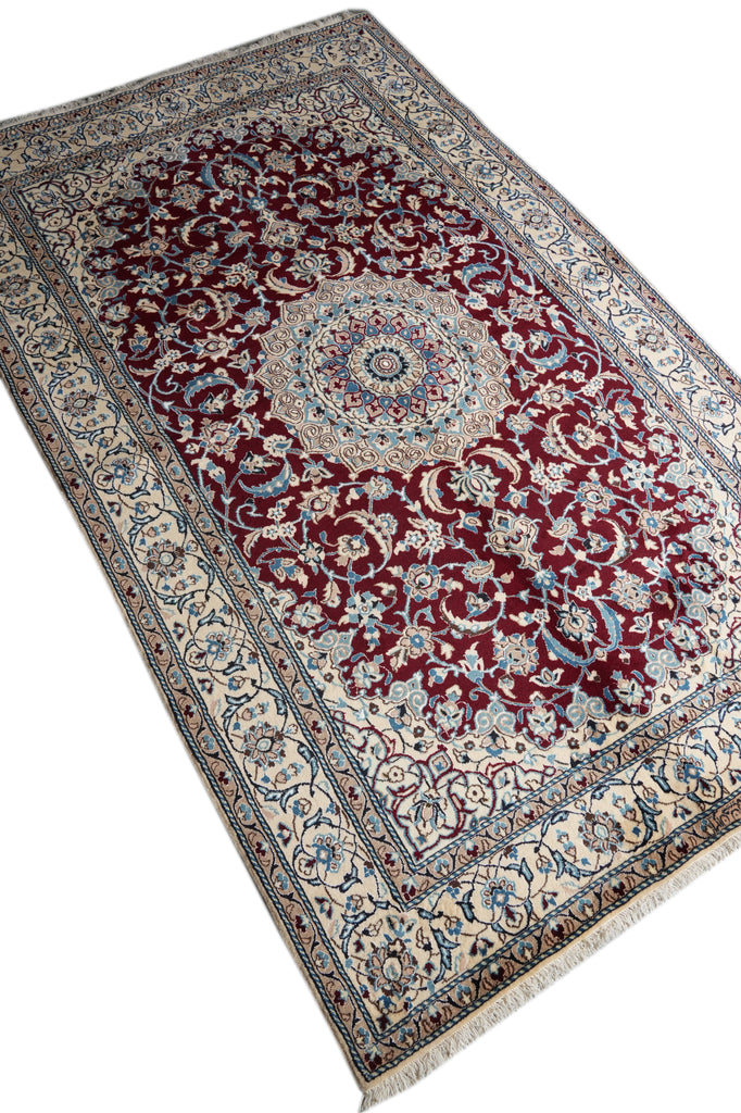 A traditional Persian-style area rug with an intricate floral pattern, featuring a deep red central field, cream borders, and blue accents, shown on a plain white background.