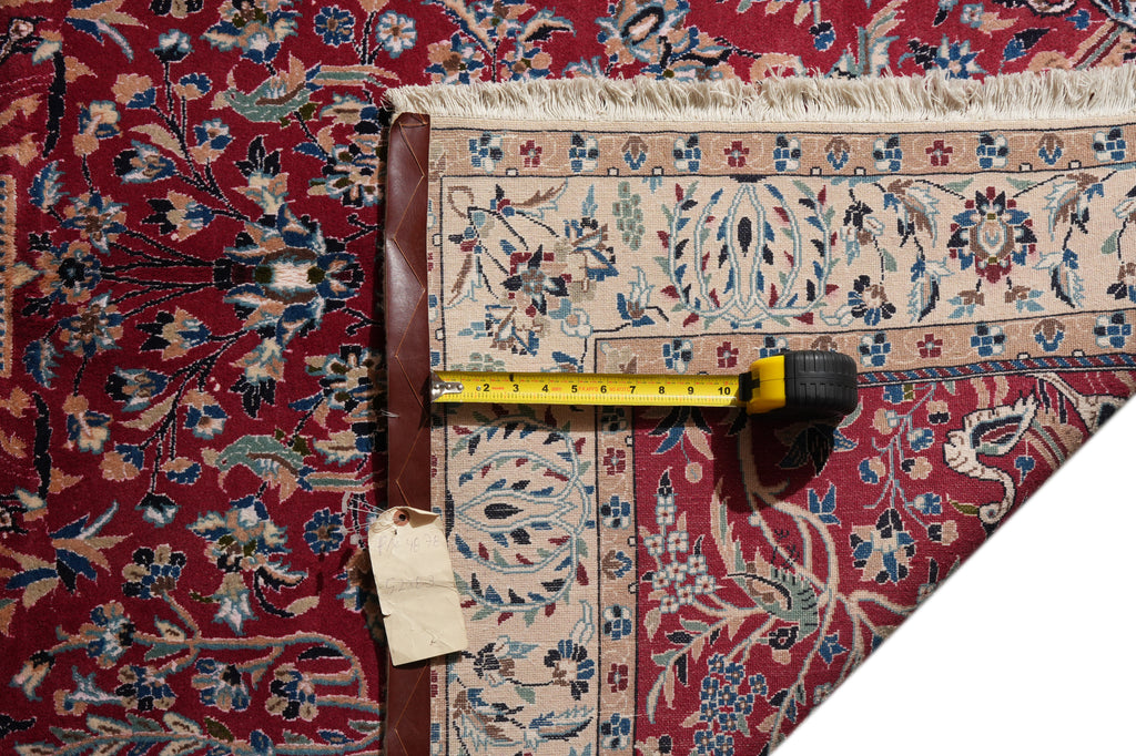 A close-up of a patterned area rug corner with a tape measure laid across it to show dimensions. The rug has a deep red field with cream, blue, and brown floral motifs and a decorative border.
