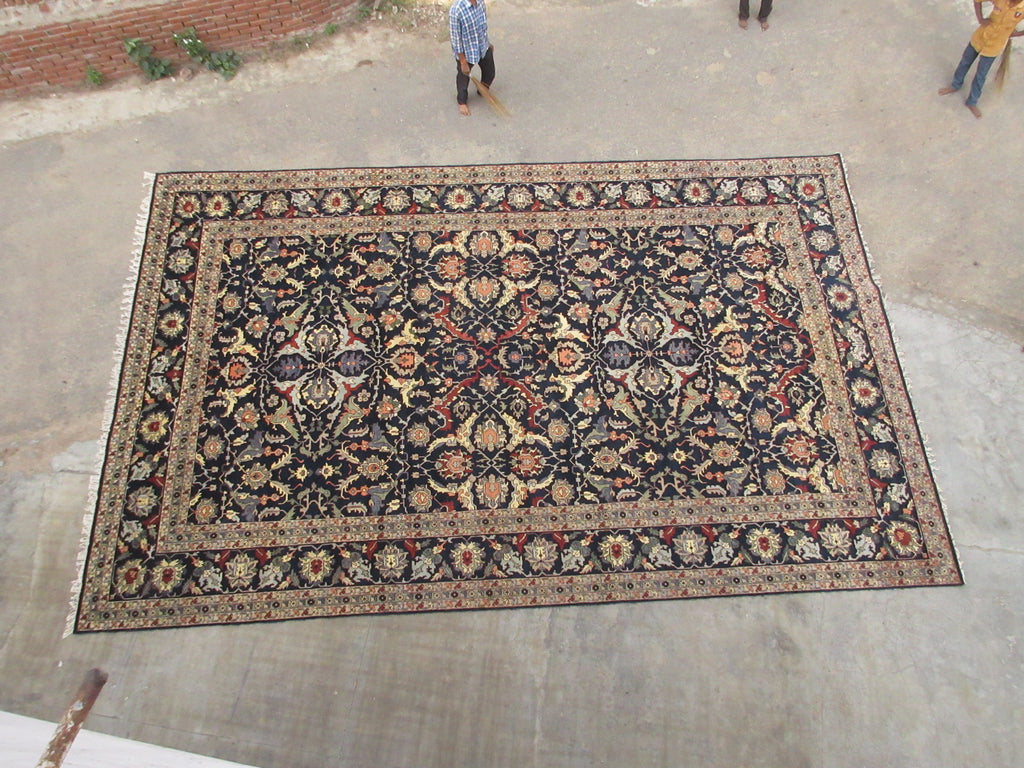A large rectangular traditional area rug with an intricate floral and geometric pattern in beige, black, red, and blue, laid out on a concrete floor .