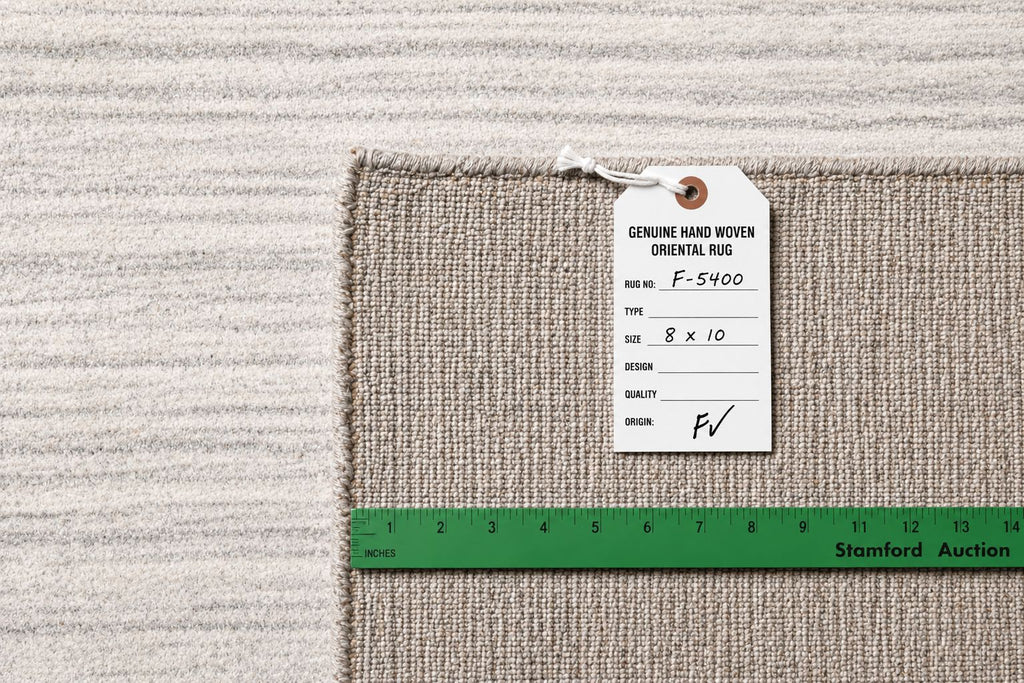 A close-up of a woven rug corner with a white product tag attached and a green measuring ruler along the edge, showing the rug texture and dimensions against a matching rug background.