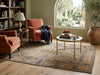 Load image into Gallery viewer, A styled living room scene featuring a large traditional oriental-style area rug in beige, blue, and rust tones. The rug is placed on a light wood floor, surrounded by two rust-orange armchairs, a round white coffee table with gold legs, a bookshelf, and a large potted olive tree by an arched window.