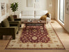 Load image into Gallery viewer, A large traditional-style area rug with a burgundy and beige floral pattern is shown in a modern living room setting with a white sofa, olive green armchairs, a wooden coffee table, and a potted plant.