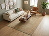 Load image into Gallery viewer, A styled living room scene showing a large rectangular area rug with an ornate vintage floral pattern in muted beige, sage green, and terracotta tones. The rug is placed on a light wood floor beneath a cream sofa, a dark wood coffee table, a brown leather armchair, and a potted plant.