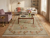 Load image into Gallery viewer, A large rectangular area rug with a floral border pattern in muted beige, sage green, and peach tones, styled in a bright living room with a wooden coffee table, sofa, and armchair.