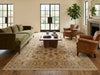 Load image into Gallery viewer, A large traditional oriental area rug in beige, blue, and brown tones is styled in a bright living room with a green sofa, brown leather armchairs, a wooden coffee table, and large black-framed windows.
